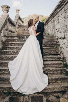 Gorgeous wedding couple embracing on stone stairs near old castle in park. Stylish beautiful bride in amazing gown and groom kissing on background of ancient building